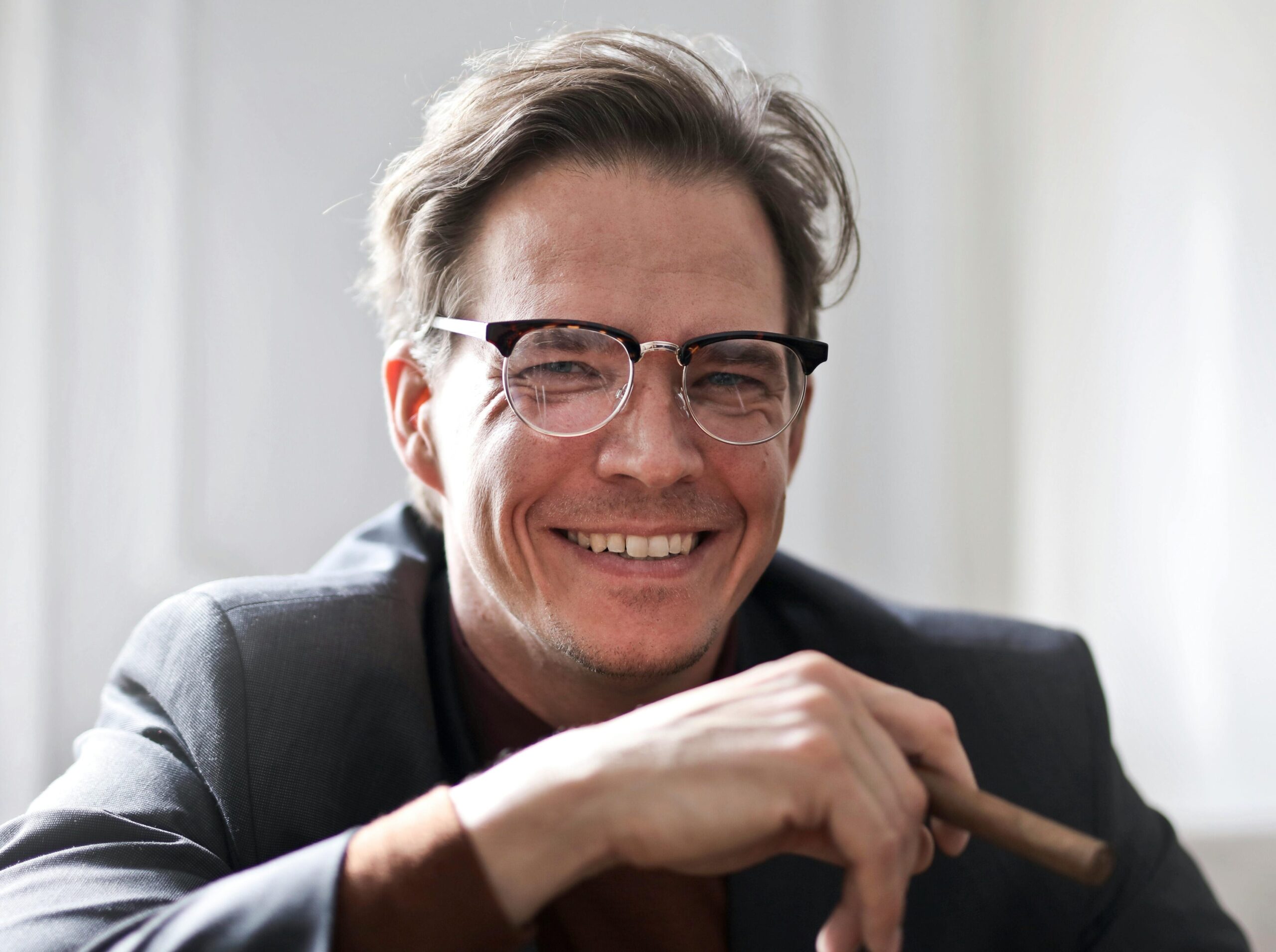 Cheerful businessman in glasses and suit holding a cigar, indoors.